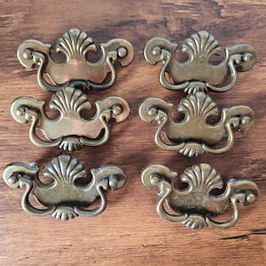 May include: Six brass-toned metal drawer pulls arranged in two columns. Each pull features a central shell-like motif at the top and bottom, with curved, scrolling arms extending to the sides. The hardware has a weathered, antique finish.