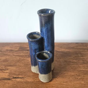 May include: A handmade ceramic vase with three cylindrical openings. The vase is primarily a deep blue color with a lighter blue gradient and a tan base. The vase is displayed on a wooden surface.