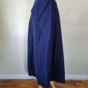 May include: A long, navy blue skirt with a button-up front and decorative stitching along the hem. The skirt is made of a solid, dark blue fabric and features several buttons down the side. The skirt is displayed on a wooden stand.