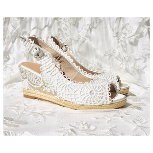 Op de afbeelding: Witte espadrilles met een peeptoe en een enkelbandje. De sandalen hebben een jute zool en een delicaat bloemenpatroon.