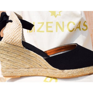 Peut inclure: Sandales compens&eacute;es en espadrille noires avec une plateforme en jute et un lacet noir. Les sandales sont sur une surface blanche avec le texte "Bizencas" visible.
