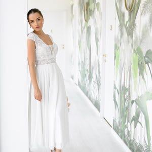 May include: A woman in a white lace wedding dress with a flowing skirt stands in a hallway with white walls and a tropical print wallpaper. She is wearing white sneakers.
