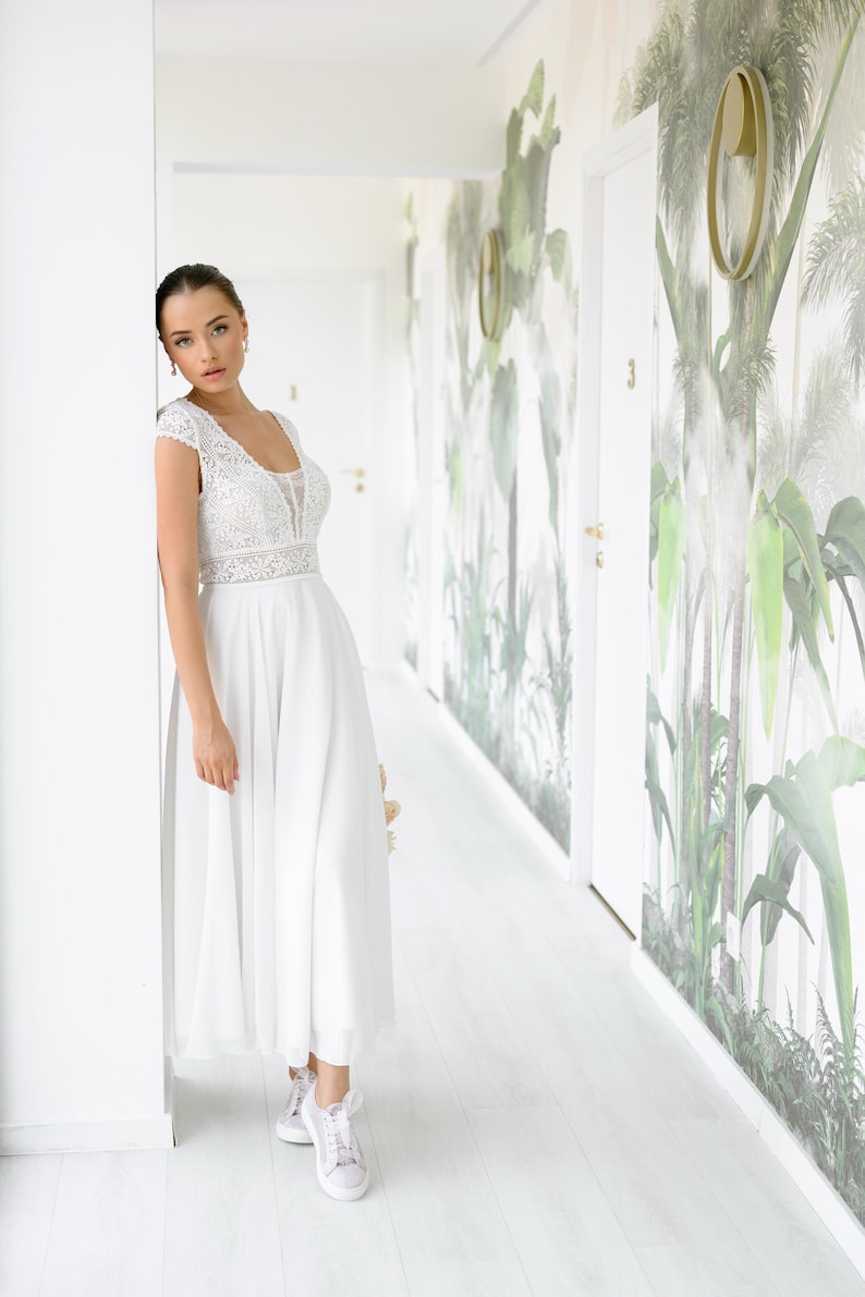 May include: A woman in a white lace wedding dress with a flowing skirt stands in a hallway with white walls and a tropical print wallpaper. She is wearing white sneakers.