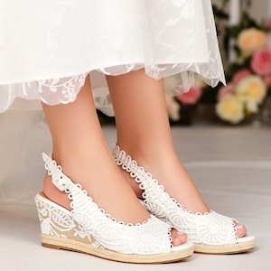 Op de afbeelding: Witte kanten sleehak sandalen met open tenen en een geweven jute zool. De sandalen hebben een delicaat bloemen kanten ontwerp en een slingback bandje. De sleehak biedt een comfortabele lift. Perfect voor een bruiloft of speciale gelegenheid.