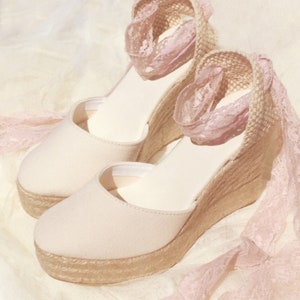 May include: A pair of beige espadrille wedge sandals with pink lace trim. The sandals have a closed toe and a thick wedge heel.