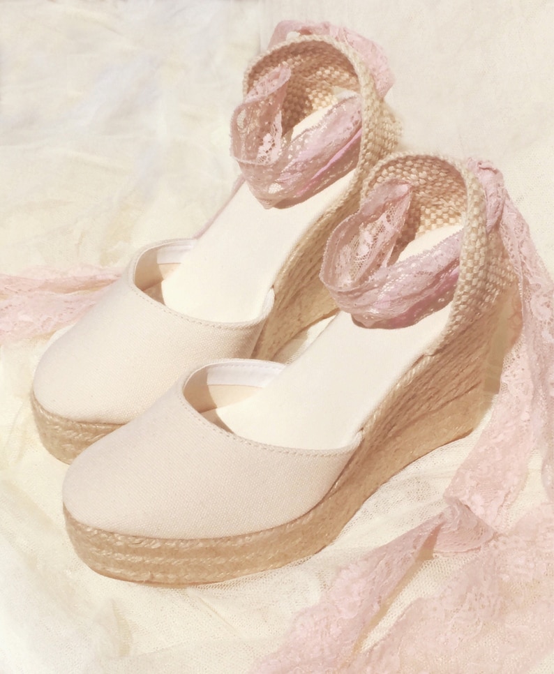 May include: A pair of beige espadrille wedge sandals with pink lace trim. The sandals have a closed toe and a thick wedge heel.