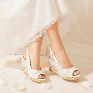 Op de afbeelding: Witte open-teen wigzolen met gedetailleerde kanten patronen. De sandalen hebben een geweven jute zool en een slingback bandje. Deze schoenen maken deel uit van een bruidscollectie, geschikt voor een bruiloft.
