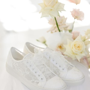 May include: A pair of white lace sneakers with a bow on the laces. The sneakers are sitting on a white surface with white flowers in the background.