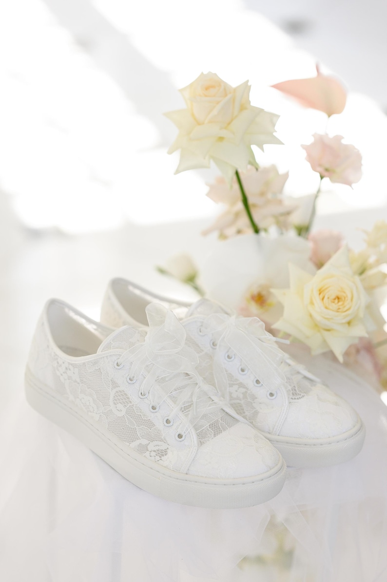 May include: A pair of white lace sneakers with a bow on the laces. The sneakers are sitting on a white surface with white flowers in the background.