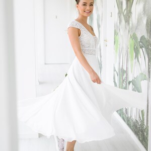May include: A woman wearing a white lace wedding dress with a flowing skirt and a fitted bodice. She is smiling and wearing white lace sneakers.