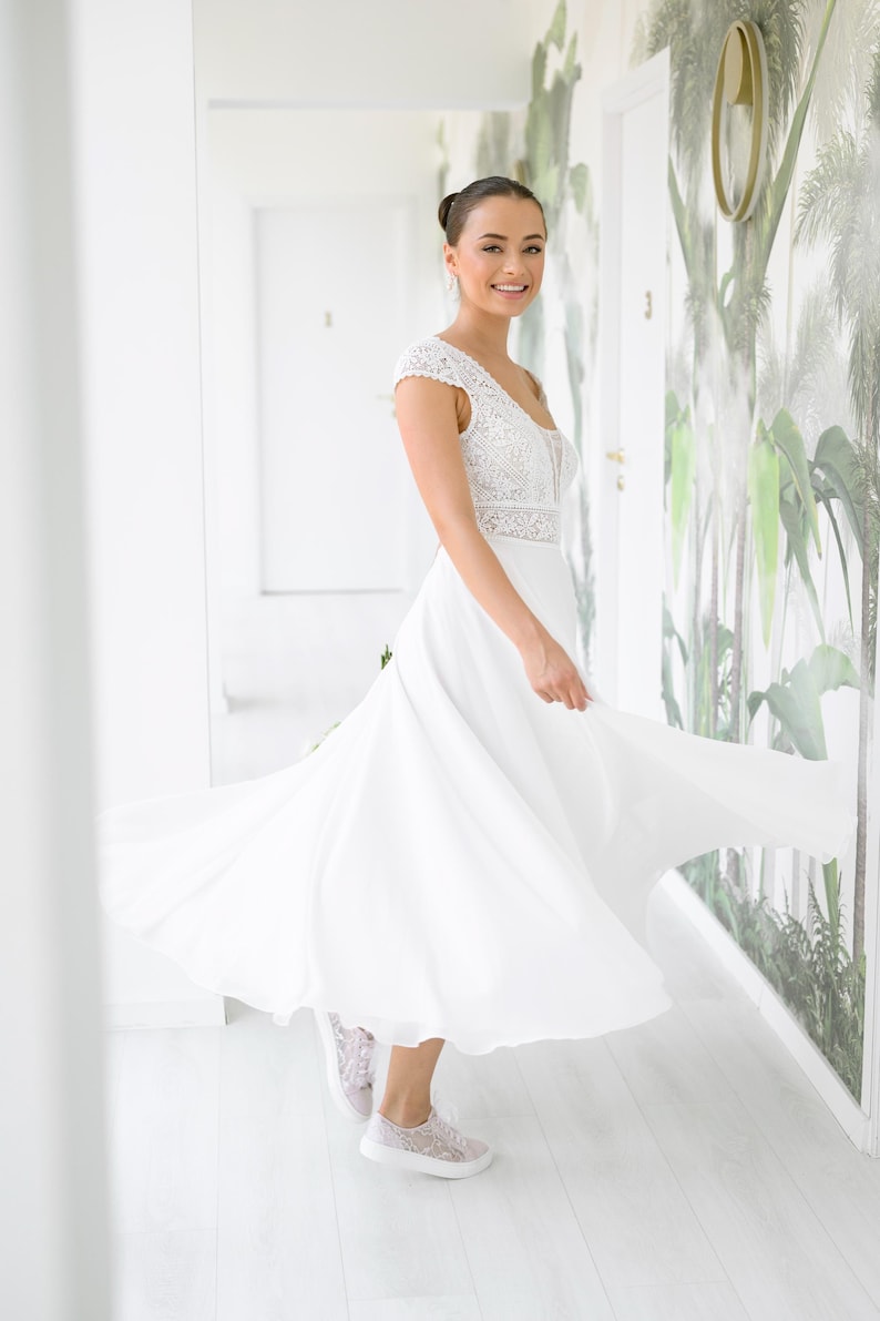 May include: A woman wearing a white lace wedding dress with a flowing skirt and a fitted bodice. She is smiling and wearing white lace sneakers.