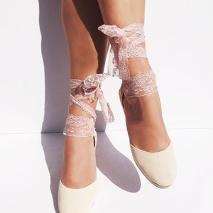 May include: A pair of cream-colored espadrille wedges with pink lace ties. The lace ties are tied around the ankles and have a bow at the top.