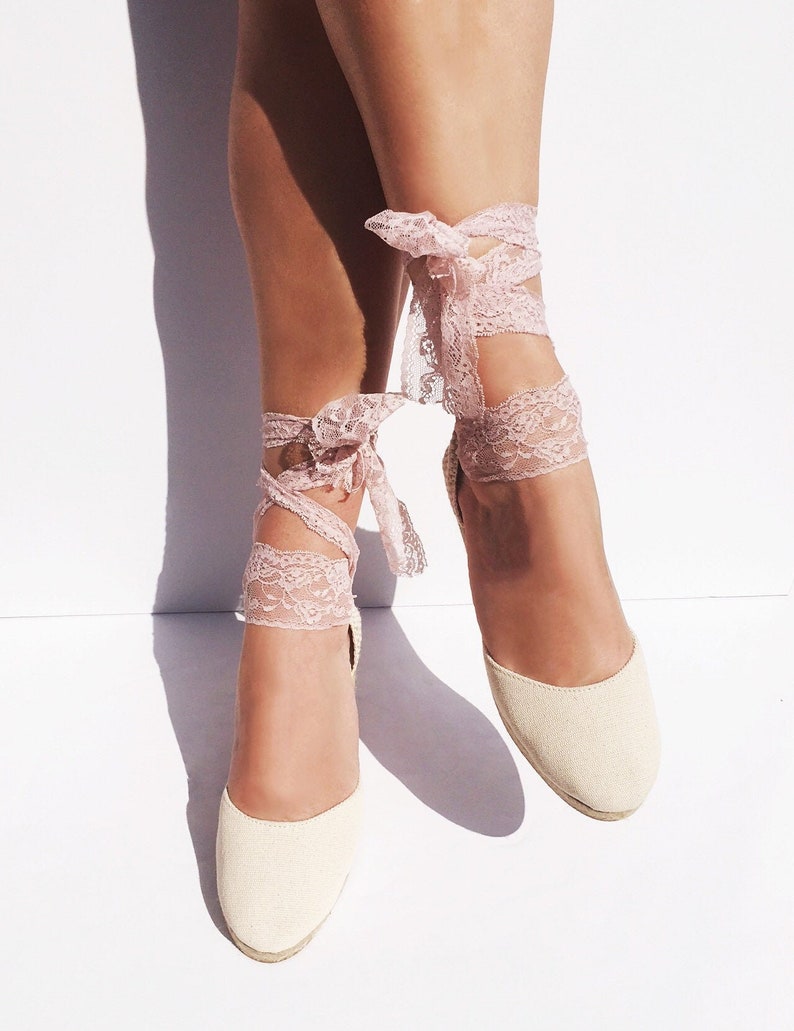 May include: A pair of cream-colored espadrille wedges with pink lace ties. The lace ties are tied around the ankles and have a bow at the top.