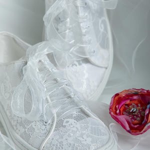 May include: A pair of white lace sneakers with satin ribbon laces. The sneakers are tied in a bow and are sitting on a white surface.