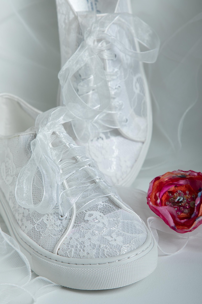 May include: A pair of white lace sneakers with satin ribbon laces. The sneakers are tied in a bow and are sitting on a white surface.