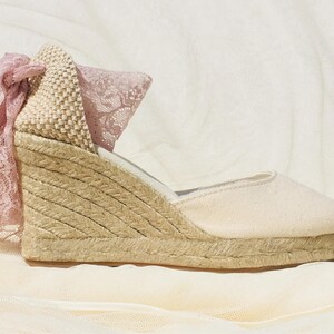 May include: A pair of beige espadrille wedge sandals with a pink lace bow detail. The sandals have a woven jute platform and a canvas upper.