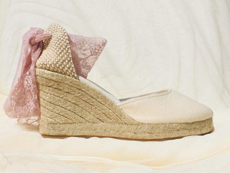 May include: A pair of beige espadrille wedge sandals with a pink lace bow detail. The sandals have a woven jute platform and a canvas upper.