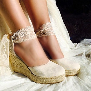 May include: A pair of off-white espadrille wedge sandals with lace trim around the ankles. The sandals are on a white fabric surface.