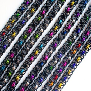 May include: Multiple black chainmail bracelets with colorful accents. The bracelets are arranged in a diagonal line on a white background.