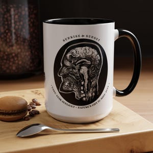 May include: White ceramic mug with a black rim and handle. The mug features a black and white illustration of a human head with internal organs visible. The text "Sunrise & Sunset" is printed above the illustration and "DAWN - LUSK SUNLIGHT - NATURE'S GIFT TO MIND & BODY" is printed below the illustration.