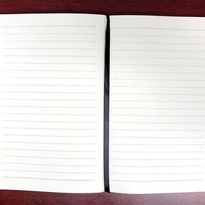 Puede incluir: Un cuaderno blanco con una encuadernaci&oacute;n negra. El cuaderno est&aacute; abierto y revela dos p&aacute;ginas en blanco con l&iacute;neas.
