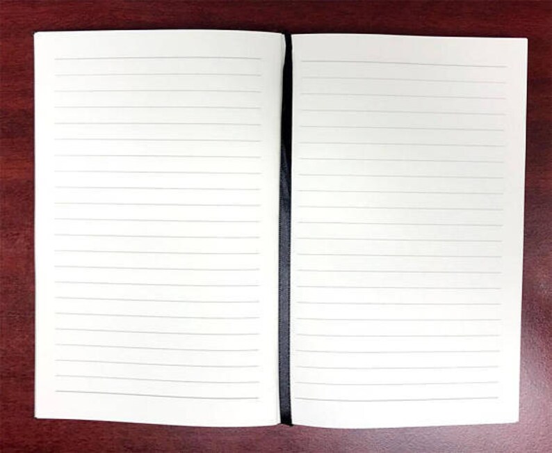 Puede incluir: Un cuaderno blanco con una encuadernaci&oacute;n negra. El cuaderno est&aacute; abierto y revela dos p&aacute;ginas en blanco con l&iacute;neas.