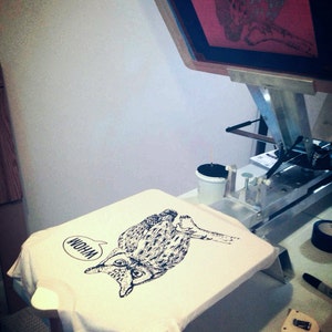May include: A white t-shirt with a black and white illustration of an owl with the word "Whoa" in a speech bubble above it. The t-shirt is being printed on a screen printing press.