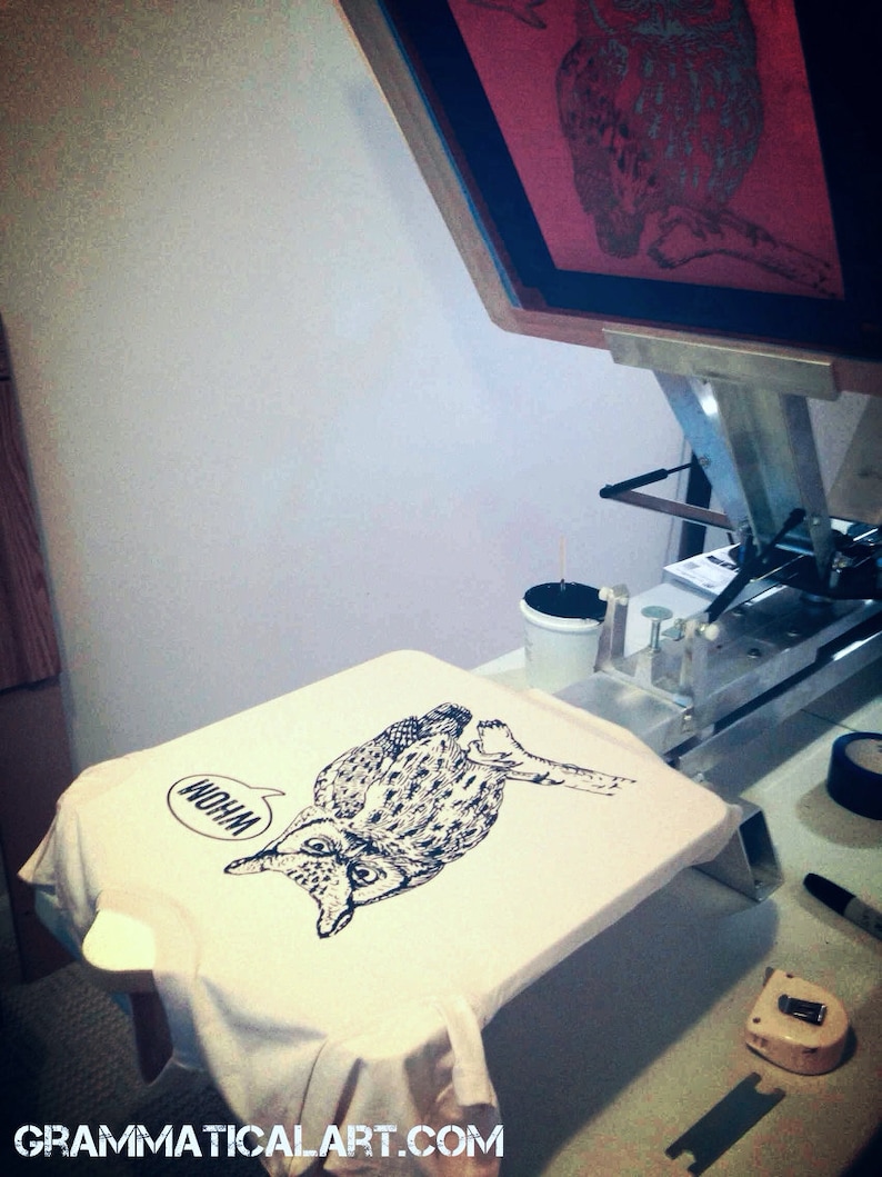 May include: A white t-shirt with a black and white illustration of an owl with the word "Whoa" in a speech bubble above it. The t-shirt is being printed on a screen printing press.