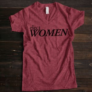 May include: A maroon v-neck t-shirt with the text "elect WOMEN" printed in black.