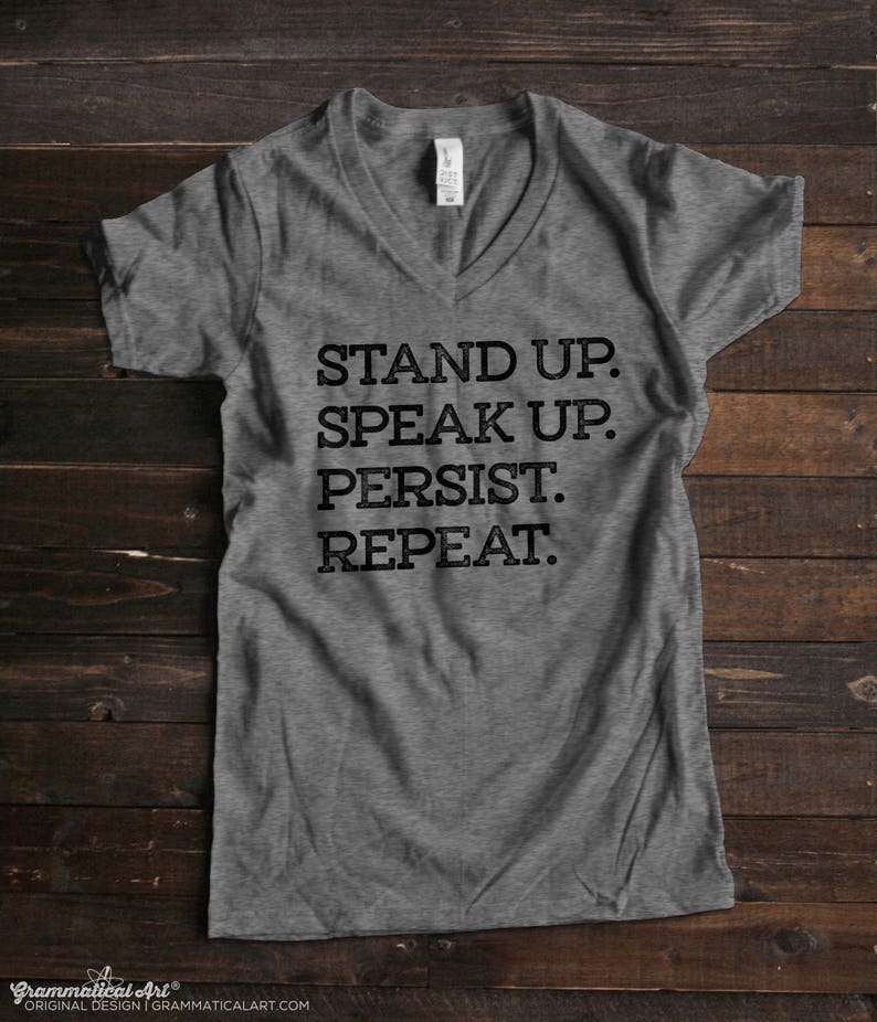 May include: Gray V-neck t-shirt with the text "STAND UP. SPEAK UP. PERSIST. REPEAT."