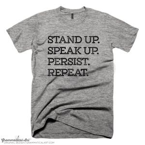 May include: Gray t-shirt with the text "STAND UP. SPEAK UP. PERSIST. REPEAT."