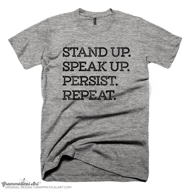 May include: Gray t-shirt with the text "STAND UP. SPEAK UP. PERSIST. REPEAT."
