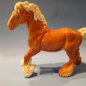 May include: A ceramic horse figurine in shades of brown with a cream-coloured mane, tail, and fetlocks. The horse is depicted in a walking pose. The figurine has a glossy finish and is set against a blue backdrop.
