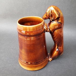 May include: Brown ceramic beer stein with a dachshund dog handle. The mug has a ribbed design and a glossy finish. The dog handle is sculpted in a standing position, appearing to hold the mug.