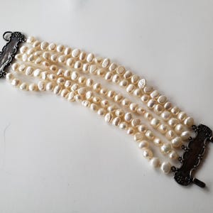 May include: A multi-strand bracelet featuring creamy white pearls. The bracelet has five strands of irregularly shaped pearls, secured by ornate, dark metal clasps. The pearls have a natural luster, and the bracelet lies on a white surface.