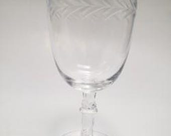 May include: A clear wine glass with a decorative etched leaf pattern around the top. The glass has a round base and a detailed stem. The glass is empty and sits on a white surface.