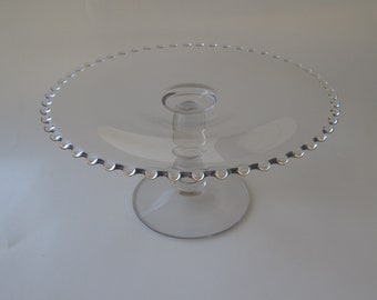 May include: A clear glass cake stand with a round, beaded edge. The stand has a short, cylindrical stem and a circular base. The design is simple and elegant, suitable for displaying cakes or other desserts.