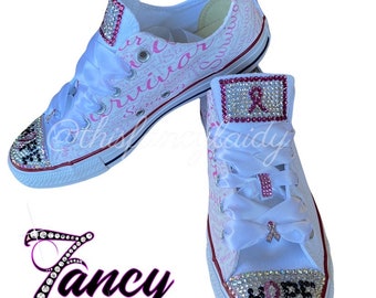 Könnte beinhalten: Weiße Canvas-Sneaker mit rosa Schleifen und Strass-Akzenten. Die Schuhe sind mit dem Wort "HOPE" verziert und die Worte "SURVIVOR" sind an der Seite aufgedruckt.