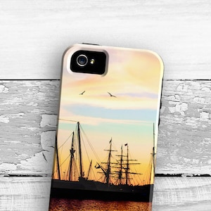 May include: A smartphone case featuring a sunset scene with a silhouette of a tall ship on the water. The sky transitions from yellow to blue, with two birds in flight. The case has the text "Lil Bits Of Light" at the bottom.