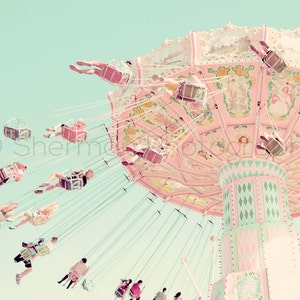 May include: A vintage-style carousel with pink and green paint, featuring a whimsical design. The carousel is in motion, with people riding in the swings. The background is a soft blue.