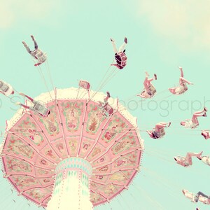 May include: A pink and white carousel with people riding in swings against a light blue sky. The carousel has a colorful, ornate design with a central pole and a large, circular platform.
