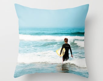 May include: Cushion depicting a surfer in a black wetsuit entering the sea with a yellow and white surfboard. The background shows a blue sky and ocean waves, creating a coastal theme.