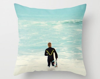 May include: A person in a black wetsuit walks towards the shore with a surfboard under their arm. The ocean is a light blue colour with white waves.