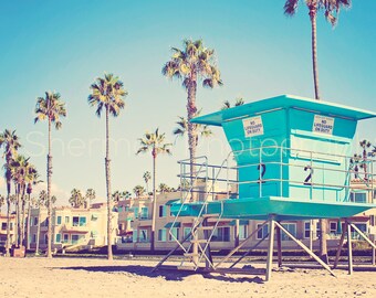 Könnte beinhalten: Ein türkisfarbener Rettungsschwimmerturm mit einem weißen Schild mit der Aufschrift "No Lifeguard on Duty" an einem Strand mit Palmen und einer Reihe von Häusern im Hintergrund.