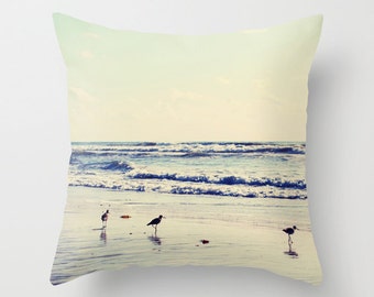 May include: A beach scene with three birds walking on the sand. The birds are silhouetted against the blue ocean and white waves. The sky is a light blue with white clouds.