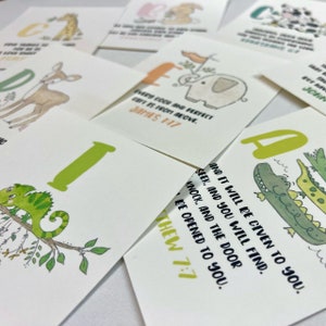 Op de afbeelding: Een set alfabetkaarten met kleurrijke illustraties van dieren en een bijbelvers op elke kaart. De kaarten tonen een kameleon, een giraffe, een hert, een olifant, een krokodil en een konijn. De verzen zijn "Elke goede en volmaakte gave komt van boven. Jakobus 1:17" en "Zoek, en je zult vinden; klop, en er zal voor je worden opengedaan. Matthe&uuml;s 7:7."