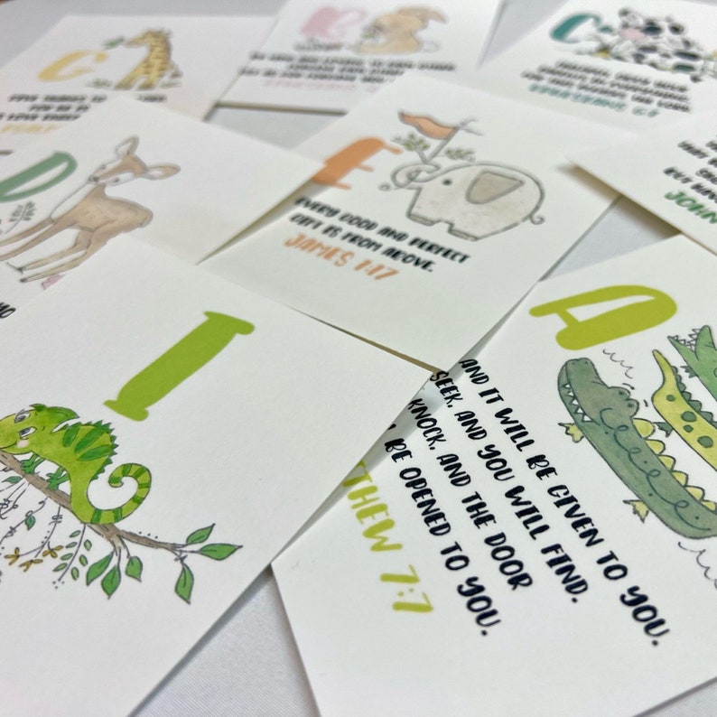 Op de afbeelding: Een set alfabetkaarten met kleurrijke illustraties van dieren en een bijbelvers op elke kaart. De kaarten tonen een kameleon, een giraffe, een hert, een olifant, een krokodil en een konijn. De verzen zijn "Elke goede en volmaakte gave komt van boven. Jakobus 1:17" en "Zoek, en je zult vinden; klop, en er zal voor je worden opengedaan. Matthe&uuml;s 7:7."