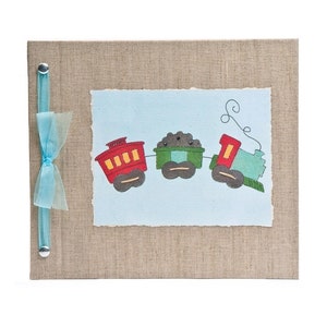May include: A handmade scrapbook with a tan linen cover and a light blue ribbon closure. The center features a light blue paper with a colorful train illustration. The train cars are red, green, and blue.