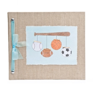May include: A burlap-covered photo album with a blue ribbon and silver metal accents. The front cover features a hand-painted illustration of a baseball bat with a baseball, basketball, football, and soccer ball hanging from it.