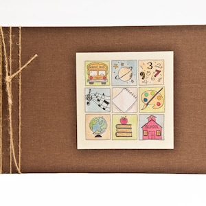May include: A brown fabric-covered scrapbook with a beige paper insert featuring a colorful illustration of school-related items, including a school bus, a globe, a notebook, a paint palette, and a schoolhouse. The text "School Bus" and "School" are visible on the illustration.